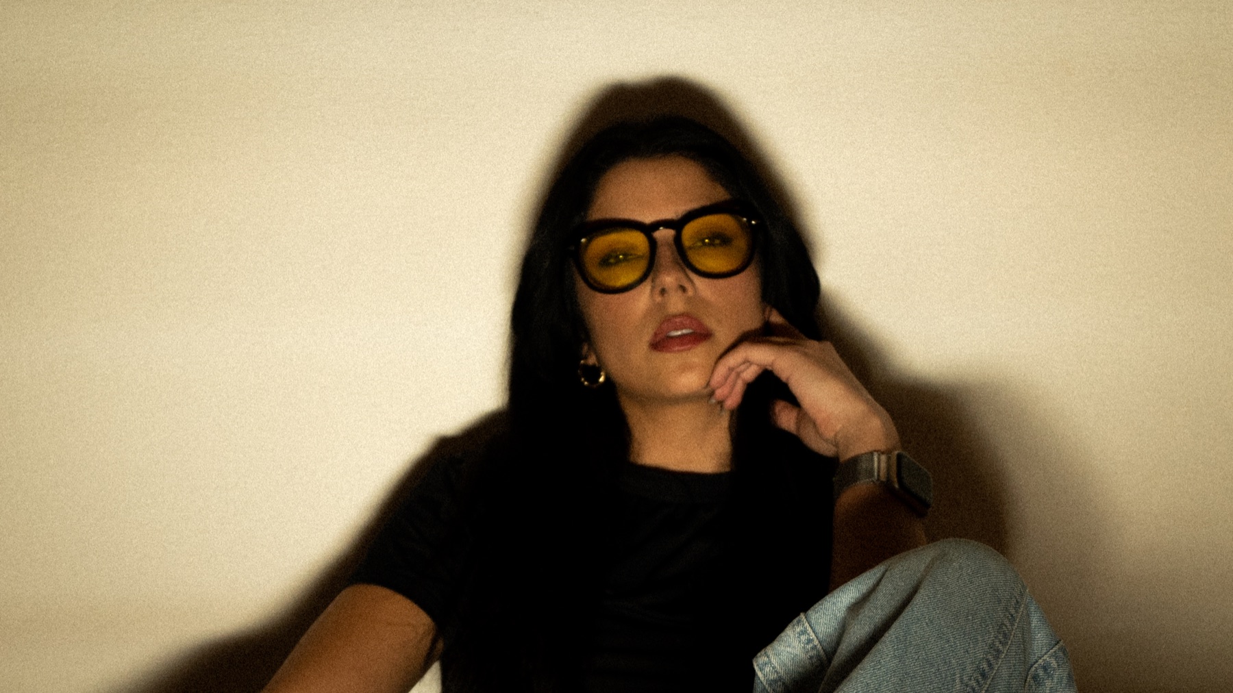 Janis Jane, lunettes ambrées et tenue noire, assise devant un mur clair